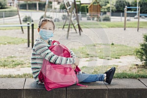 A girl in a protective mask sits in the schoolyard with her pink backpack. Rest from study. Keeping distance