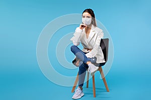 A girl in a protective mask and a white jacket sits on an office chair. Blue background, empty space and the fight against the