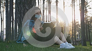 Girl in protective medical mask relaxing and reading book while sitting on grass in public park at beautiful sunset. Portrait of c
