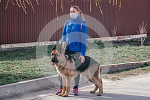 A girl in a protective medical mask walks a dog on the street. Leisure with a pet during quarantine. Walk with a German shepherd