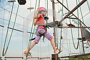 The girl in protective and safety clothing passes over a hanging bridge in a sports park, holding the side ropes