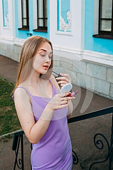 Girl in a purple dress paints her lips with lipstick, near the blue facade of a beautiful old building