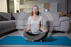 A girl in a purple scarf and in sportswear sits in a lotus pose on a yoga mat