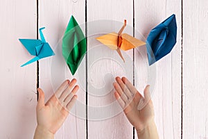 A girl puts together an origami crane and a boat made of colored paper. On a wooden background