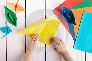 A girl puts together an origami crane and a boat made of colored paper. On a wooden background