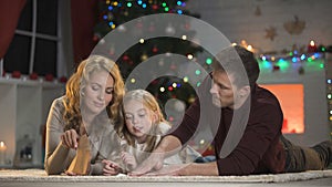 Girl putting letter for Santa into envelope, lying under X-mas tree with parents