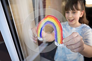 Girl Putting Picture Of Rainbow In Window At Home During Coronavirus Pandemic To Entertain Children