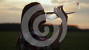 Girl quenches her thirst in a field at sunset. Silhouette of a girl drinks water from a plastic bottle, sun rays