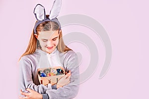 Girl in rabbit bunny ears on head with basket full of colored eggs on pink background. Cheerful smiling happy child. Easter