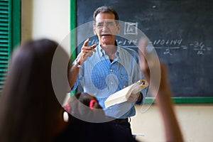 Girl Raising Hand Asking Question To Teacher At School