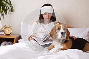 A girl is reading a book in bed under a blanket. A beagle dog is nearby.