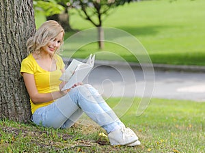 Girl reading the book. Blonde beautiful young woman with book sitting on the grass and leaning to the tree. Outdoor.