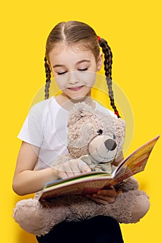 A girl is reading a book and holding a teddy bear on a yellow background