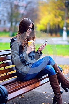 Girl reads sms, rewritten smartphone outdoors in spring or autumn, on the bench, with coffee tea, breakfast, concep