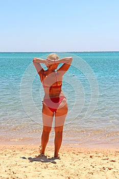 Girl in red bathing suit sunbathing standing on beach by sea. Beautiful woman