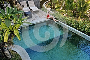 Girl in red bikini sitting on the edge of pool in jungle admires a beautiful view of the palm trees.Luxury holiday on