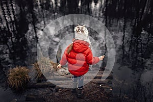 Girl in red coat staying on the bank of the lake alone and looking at reflection in water