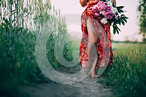 A girl in a red dress with a bouquet of peonies is walking along the path barefoot. Soft focus