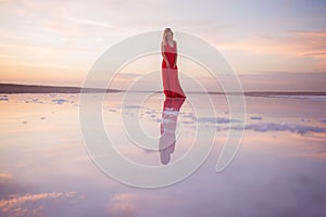 A girl in a red dress stands in the water of a mirror lake at sunset. Reflection of a girl and clouds at sunset