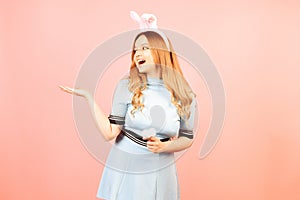 A girl with red hair in a blue dress and easter bunny ears on a pink background. A beautiful woman is pointing at a product or