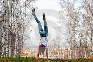 A girl with red hair stands on her hands with her back to the camera on a hill against the background of a blurred birch forest.