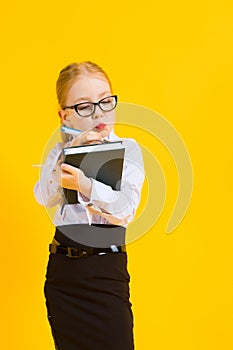 Girl with red hair on a yellow background. A charming girl in transparent glasses writes a pen in her notebook.