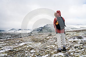 Girl with red hooded jacket and backpack are hiking in snow covered mountains with beautiful scenic landscape view and foggy weath