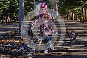 Girl in a red jacket, hat and glasses chasing pigeons in the park.