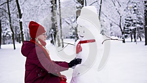 A girl in a red jacket makes a snowman in a snowy forest and knits a red scarf for him.