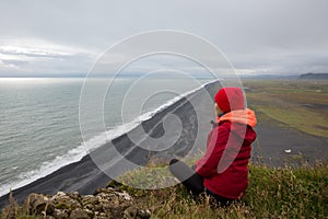 A girl in a red jacket sits on a cliff above the sea shore with black lava sand stretching to the horizon