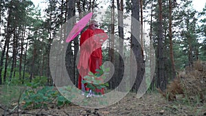 A girl in a red kimono under an umbrella walks the coniferous forest, turns back and covers her face with a fan, slow