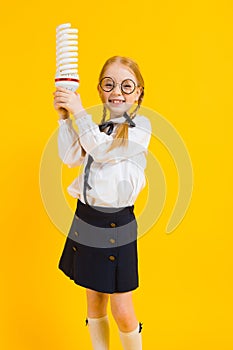 Girl with red pigtails on a yellow background. A charming girl in round transparent glasses is holding a light bulb in