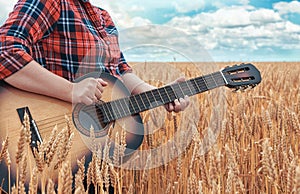 Girl in the red shirt in wheat field plays the acoustic guitar. Beautiful nature at bright sunny summer day.