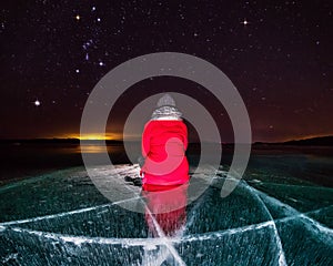 girl in red sits on an ice-lit frozen Lake Baikal under a starry night sky with the constellation Orion, Siberia, Russia