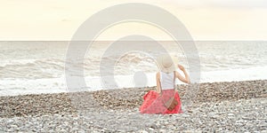 Girl in red skirt and hat sits on the seashore. Sunset time. Back view