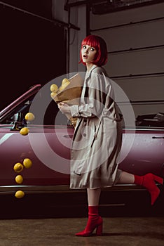 girl in red wig and stylish trench coat holding paper bag with falling lemons near retro