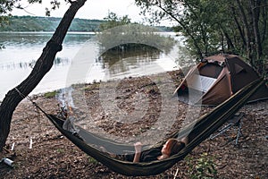 Girl relaxes in a hammock. Quickly folding tent and campfire. River bank. Wild camping. Travel equipment. Vacation and travel