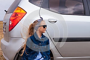 Girl relaxing and enjoying road trip sitting by the silver car. Happy child rides toward adventure. Lady in denim jacket