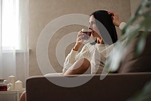 The girl is resting in a chair, drinking tea, reading a book, shopping on the phone against the background of the window