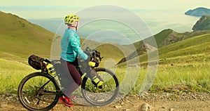 The girl rests beside her bike in mountains after her cycling journey. The woman looking at the view of beautiful lake