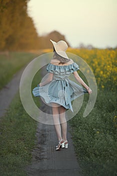 A girl in retro clothes with a straw hat runs along a rural road along a yellow field. Running away into the distance.