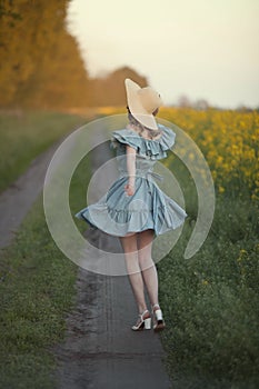 A girl in retro clothes with a straw hat runs along a rural road along a yellow field. Running away into the distance.