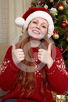 Girl in retro red sweater and santa hat show best gesture. Home interior with christmas decoration, fir tree and gifts. New year e