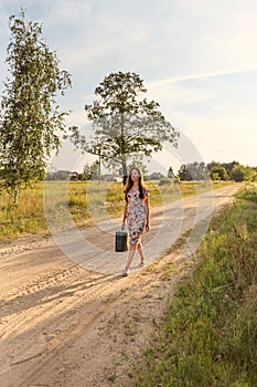 A girl in a retro vintage dress with a suitcase and long flowing hair walks along the road in the yellow rays of the sunlight