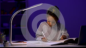 The girl rewrites the task from the textbook into a notebook. A schoolgirl sits at a table by the light of a night lamp