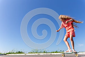 A girl is riding a skateboard with fluttering blond hair against a blue sky.