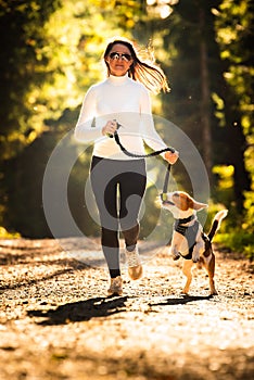 Girl is running with a dog Beagle on a leash in the autumn time, sunny day in forest. Copy space in nature