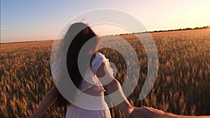 Girl runs across the field with wheat holding a man`s hand in the glare of the setting sun and laughs. Slow motion.