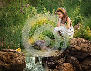 A girl in a rural dress sits near a stream and looks at a duckling