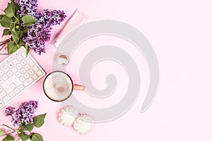 Girl`s desktop. A bouquet of lilacs, a keyboard, a cup of coffee, cakes, face cream on a pink background.